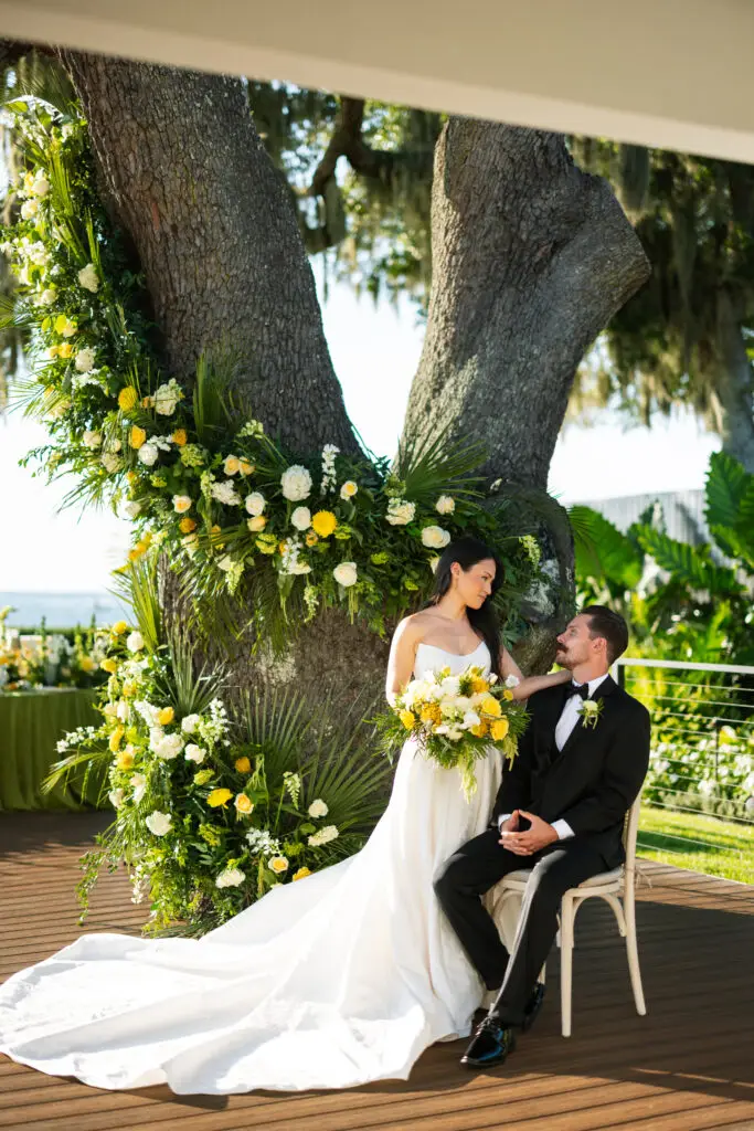 Bride and groom beneath a tree-framed yellow and white floral arch